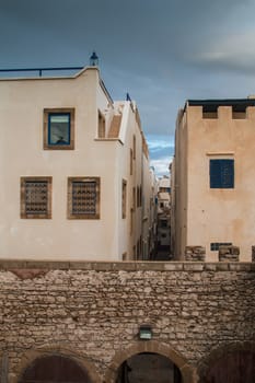 Cloudy evening sky. Wall made of stones of the city fortification. Houses with traditional architecture. Street in the middle.