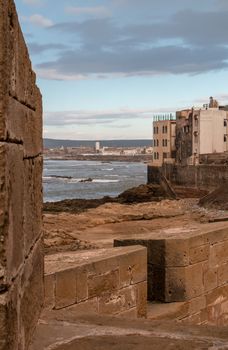 Fortification walls in the foreground. Houses of Essaouira and Atlantic ocean. Mountain on the horizon. Cloudy evening sky.