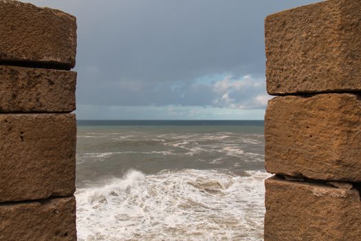 View through a window in fortification of the city Essaouira in Morocco. Rocky coastline with waves and white foam. Cloudy sky during the twilight.