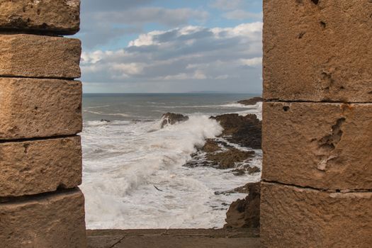 View through a window in fortification of the city Essaouira in Morocco. Rocky coastline with waves and white foam. Cloudy sky during the twilight.