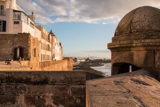 Early evening during golden hour. View on the houses on the coast in Essaouira, Morocco. Mountain on the horizon. Cloudy sky.