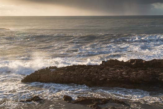 Early evening during golden hour at the Atlantic coast in Essaouira, Morocco. Big rock in the sea, intense waves with a white foam.