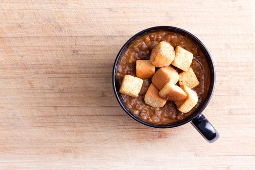 Single serving of scrumptious lentil soup garnished with toasted croutons in little bowl with handle on wooden table