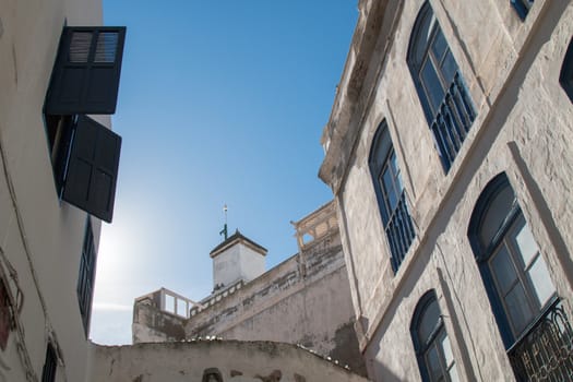 Street with high buildings with blue framed windows. In the background tower-minaret of a mosque. Bright blue morning sky. Essaouira, Morocco.