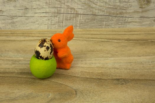 Orange Easter bunny figurine based on a green base with a little quail egg on an old wooden table. Horizontal view.