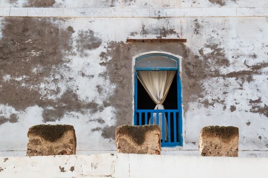 Opened window with a blue frame and grid, curtain hanging in the middle. House with a very old white damaged facade.