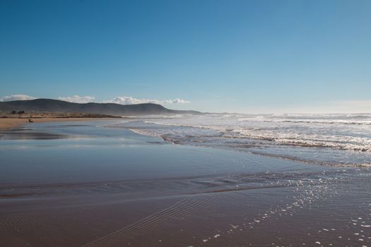 Late afternoon on the sandy beach of the Atlantic Ocean in Morocco. Waves of a tide. Mountain in the background. Blue sky with some clouds.