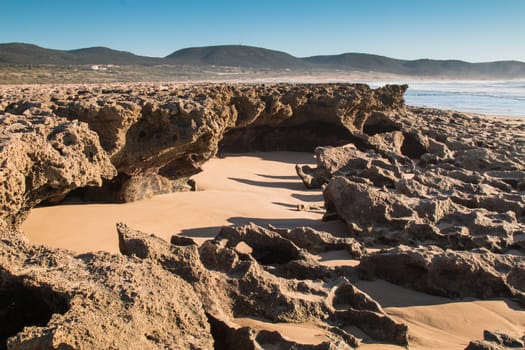 Part of the beach with a rocks at the Atlantic Ocean coast in Morocco. Hills in the background. Blue late afternoon sky.