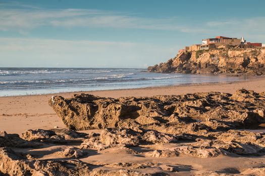 Sandy beach with rocks. Water of the Atlantic ocean. Hill with a building in the background. Cloudy sky.