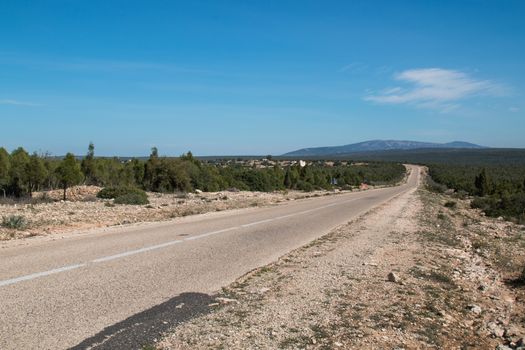 Empty country road in Morocco, lined by forests. Mountain in the background. Blue sky with a cloud.