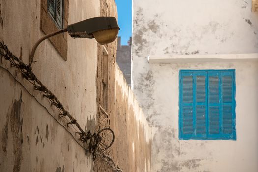 View on the corner of old houses in the city Essaouira in Morocco. Old wall, enlightened by sunshine and a window with blue shutter. Retro styled street lamp.