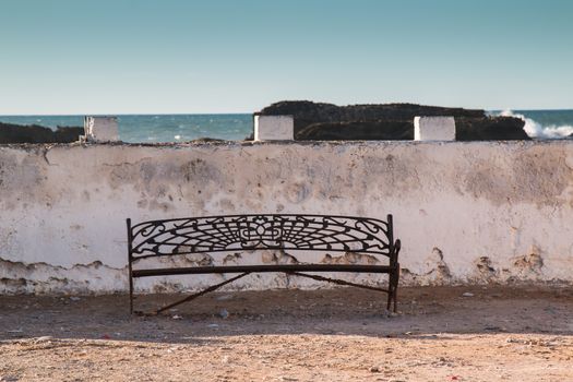 Empty iron designed bench on the banks of Atlantic ocean. Old white wall in the background, rocks and ocean. Blue early evening sky.