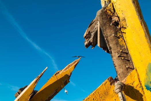 Detail of a boat wreck. Yellow painted, broken wood, nails and rope. Blue sky with trail and seagulls.