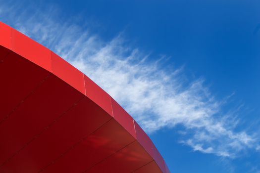 Part of a bright red modern roof with a bright blue sky in the background. Cloud lining the shape of the roof.