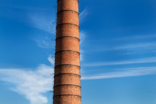 Factory high chimney, made of bricks. Blue sky in the background, with a twist of clouds around the chimney.