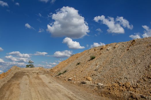 Road up the hill. Place ready for building. Devastated nature, just one tree remaining. Dry hill on the right side. Blue sky with many clouds.