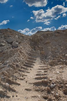 Way up the hill with a trail. Sandy soil with a lot of stones. Blue sky with many contrast clouds.