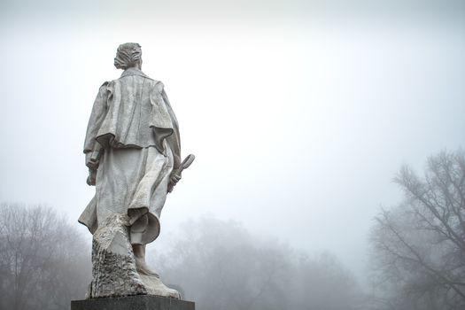 Early morning in the winter after a night rain. Intense fog. Statue of Janko Kral looking forward, trees  without leaves.