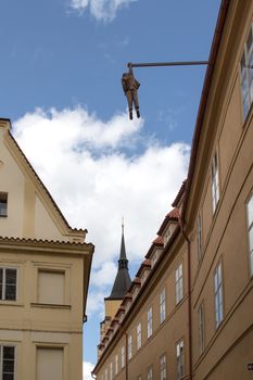 Calm street in the old city of Prague. Statue of a man, hanging above the street from the house. Old buildings, tower of a church and cloudy sky.