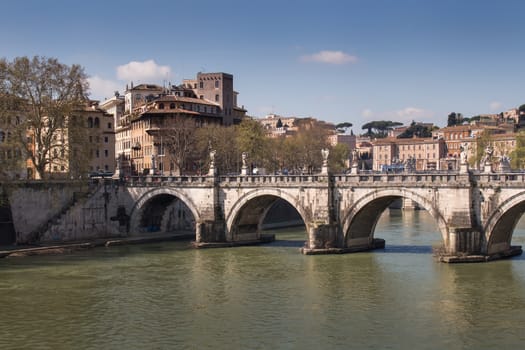 Part of Rome close to the Castle. Old bridge over the Tiber River. Buildings of the old city in the background. Blue sky with few white clouds.