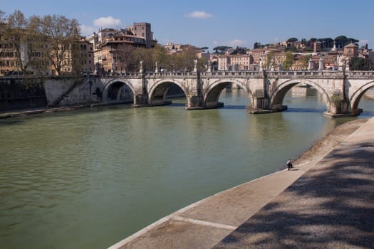 Part of Rome close to the Castle. Old bridge over the Tiber River. Buildings of the old city in the background. Blue sky with few white clouds.