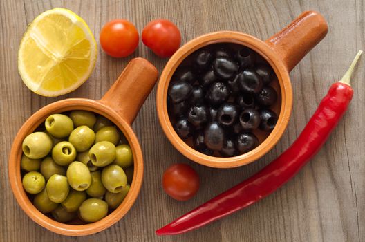 Green and black olives in a ceramic dish. Wooden table, lemon and vegetables.