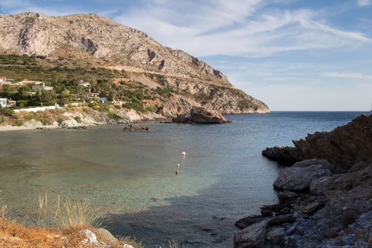 Peninsula of the greek coast. Rocky mount, partly covered with trees. Several houses. Sea bay. Clear water. Cloudy sky.