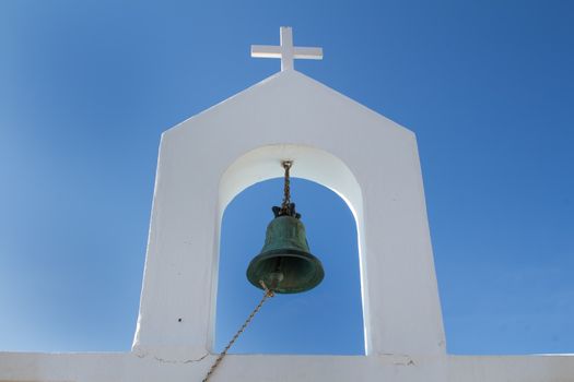 Detail of the architecture of a chapel in Greece. Heavy bell with a rope to use it. White tower with a cross. Bright blue sky in the background.