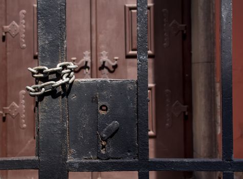 Grid fence with a missing handle. Twisted chain to keep it closed. Massive wooden decorated gate in the background.