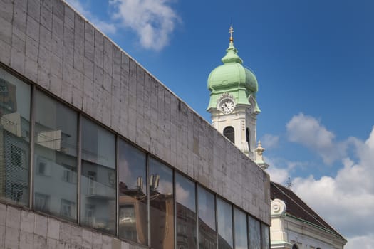 Contrast of a modern architecture made of marble and glass with a tower of baroque church in the background. Blue sky with white clouds.