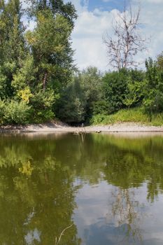 Summer cloudy day. View on a small river, having a green color from the reflection of the forest, which is lining it.