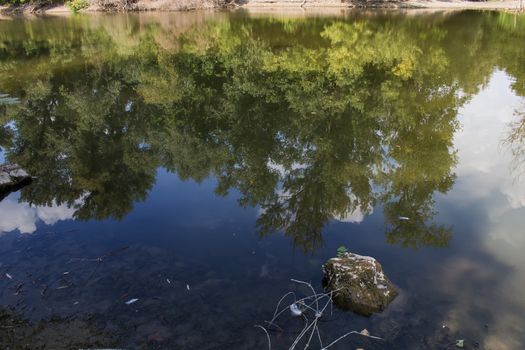 Calm summer day, forest and sky reflection in the river. Big stone in the foreground.