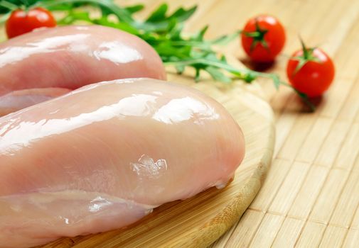 Raw chicken fillet with cherry tomatoes and arugula on a round bamboo cutting board.