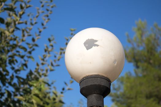 Blue summer sky. Steet lamp among the bushes. Broken white ball of the lamp. Reflection of the sun inside the ball.