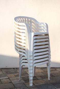 White plastic chairs strored in a column. Stone floor, white wall in the background. Outdoors. Early evening sunlight.