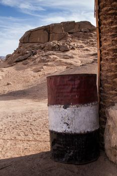 Barrel painted in national egyptian colors. Bedouin village in the desert. Rocky hill in the background. Cloudy sky.