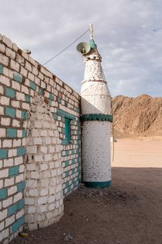 Details of the bedouin small mosque in the desert between the mountains in Egypt. Small minaret. Wall of the mosque made of contrast colors bricks. Sand everywhere around. Mountains in the background. Cloudy sky.