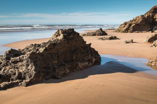 Sandy beach on the coast of Atlantic Ocean in Morocco. Wet sand, small waves, rocks in the foreground, bigger in the background. Cloudy sky.