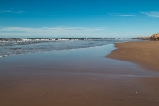 Sandy beach on the coast of Atlantic Ocean in Morocco. Wet sand, small waves, rocks in the background. Cloudy sky.