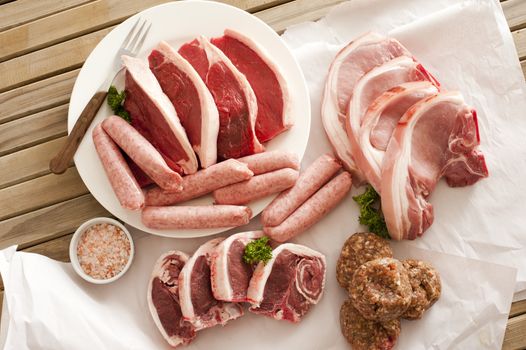 Assortment of raw meat on a wooden table with pork cutlets, lamb chops, sausage, beef steak and patties and a bowl of rock salt