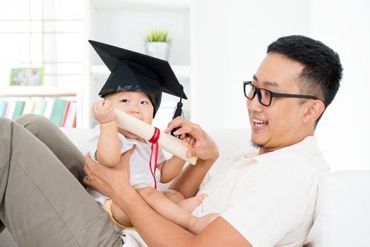 Asian family lifestyle at home. Baby with graduation cap holding certificate with father. Parent and child early education concept.