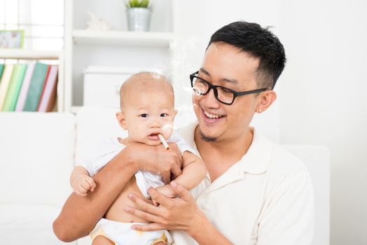 Asian family at home. Bad father giving cigarette to baby. Cigarette with lit and smoke. Unhealthy lifestyle or stop smoking concept photo.