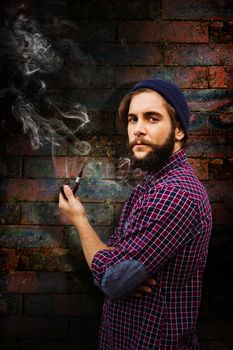 Side view of hipster holding smoking pipe against texture of bricks wall