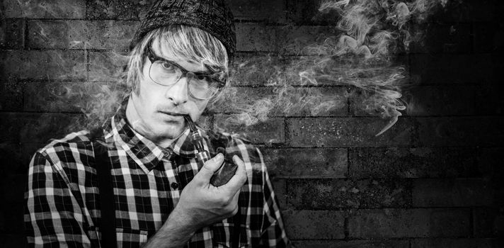 Serious blond hipster smoking a pipe against texture of bricks wall
