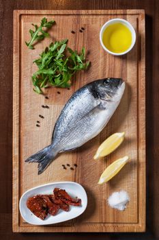 Fresh uncooked dorado or sea bream fish with lemon, herbs, oil and spices on rustic wooden cutting board, top view