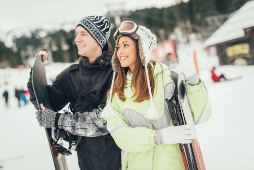 Beautiful young couple enjoying in winter vacations. They standing with skis and snowboard and looking away.