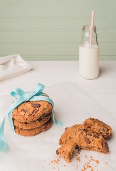 Chocolate chip cookies with a blue ribbon and a glass of milk with a straw on a white wooden table with a robin egg blue background. Vintage look.