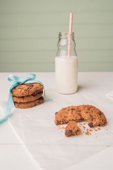 Chocolate chip cookies with a blue ribbon and a glass of milk with a straw on a white wooden table with a robin egg blue background. Vintage look.