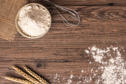 Flour in ceramic bowl with a handful with ears of wheat and beater on the wooden table closeup. Top view with copy space