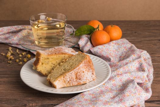 Slices of clementine cake with powdered sugar topping and cup of chamomile tea. Cake on a plate with fresh clementines on wooden board.